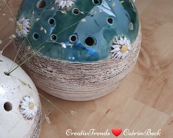 May include: A ceramic vase with a green glazed top and a white bottom. The top has a pattern of white daisies and holes for flowers. The bottom has a textured, wood-like finish.