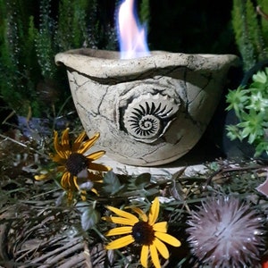 Op de afbeelding: Een witte keramische vuurkorf met een spiraalvormig ontwerp en een blauwe vlam die erin brandt. De vuurkorf staat op een bed van bruine takken en bladeren.