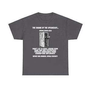 May include: Dark gray t-shirt with a graphic featuring a person in a doorway holding a bottle. Text on the shirt reads "THE SHAME OF THE SPEAKEASY... DURING MARDI GRAS" and "DON'T LET ILLEGAL LIQUOR RUIN YOUR FUN AND FESTIVITIES!"