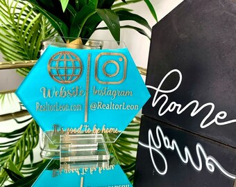 May include: Two turquoise blue hexagonal acrylic signs with gold lettering and icons. The top sign says "Website Instagram Realtorleon.com @RealtorLeon" and the bottom sign says "Realtorleon.com @Realtorleon Website Instagram". The signs are stacked on top of each other and are sitting on a clear acrylic stand.