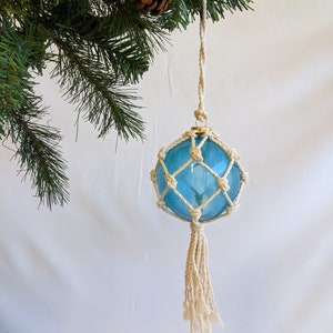 Puede incluir: Un adorno de bola de vidrio azul con una red de cuerda blanca y un borla. El adorno está colgado de una rama de un árbol de Navidad artificial verde.