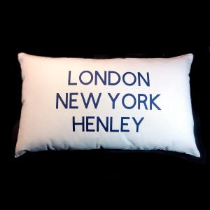 Może przedstawiać: Prostokątna biała poduszka z granatowymi napisami "LONDON", "NEW YORK" i "HENLEY". Poduszka jest na czarnym tle.