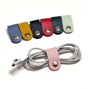May include: Assortment of seven leather cord organizers in various colors, including blue, yellow, black, red, green, light green, and pink. Each organizer has a silver button. One pink organizer is shown holding a gray cable.