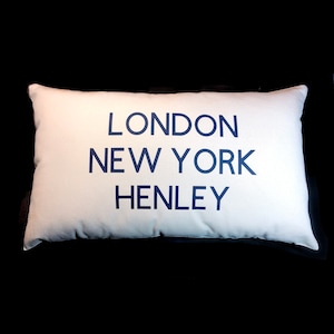 Può includere: Un cuscino rettangolare bianco con il testo "LONDON NEW YORK HENLEY" stampato in blu scuro.