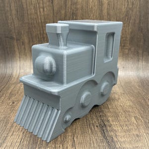 May include: A gray 3D-printed toy train. The train features a detailed design with a smokestack, a round front, and wheels. The front of the train has a series of vertical lines. The toy is made of a solid, light gray material.