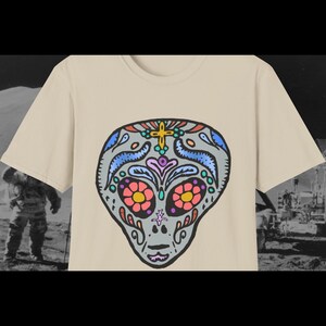 May include: A beige t-shirt with a colorful alien skull design. The skull is decorated with flowers and swirls in shades of blue, pink, yellow, and green.