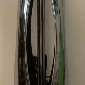 May include: A chrome table lamp with a unique, geometric design. The lamp has a cylindrical base and a tall, slender shade with a cut-out pattern. The lamp is lit and the light shines through the cut-outs, creating a decorative effect.