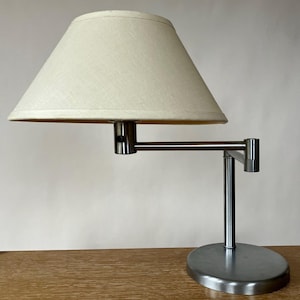 May include: A table lamp with a brushed metal base and a multi-jointed adjustable arm. The lamp features a conical, off-white fabric lampshade that provides a neutral aesthetic. The structure is composed of a vertical post and a horizontal arm.