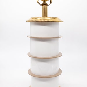 May include: A white and gold table lamp with a stacked design. The lamp has a brass base and a white cylindrical shade. The shade is made up of four stacked sections, with each section having a thin gold band.