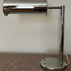 May include: A chrome desk lamp with a cylindrical shade and a round base. The lamp has a single arm that extends from the base and supports the shade.