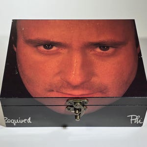 以下が含まれることがあります： 蓋に男性の顔のクローズアップ画像が描かれた黒い木製の箱。箱には真鍮製の留め金があり、箱の底には「Roquived」と「Phi」の文字が書かれている。