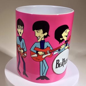 May include: A cylindrical ceramic mug with a bright pink background featuring a cartoon illustration of The Beatles. The band members are depicted playing red electric guitars and wearing light blue suits. The text The Beatles is printed in black.