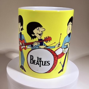 May include: A bright yellow ceramic mug featuring a colorful cartoon illustration of The Beatles. The design depicts the band members playing instruments, including a red bass guitar and a drum set with the text The Beatles printed on the front.