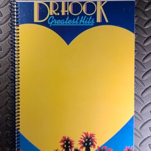 May include: A blue and yellow spiral bound notebook with a heart-shaped design. The heart is yellow and has the text "DR. HOOK Greatest Hits" in a blue and pink font.