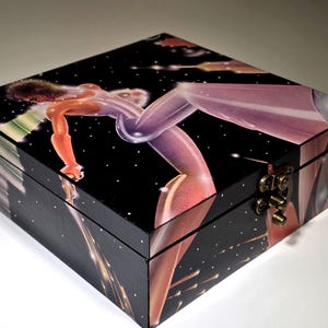 May include: A square black wooden box featuring a vibrant, airbrushed-style illustration of a figure in a space-themed setting with stars and light streaks. The box has a hinged lid and a metallic brass-colored latch on the front edge.