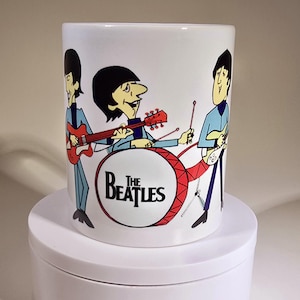 May include: White ceramic mug featuring a cartoon illustration of The Beatles band members. The design includes the band's name on a red drum and the musicians playing instruments. The mug is cylindrical and has a glossy finish.