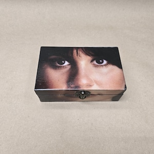 Könnte beinhalten: Eine rechteckige Holzkiste mit Scharnierdeckel und Metallverschluss. Die Oberseite der Kiste zeigt ein Nahaufnahmebild der Augen und eines Teils des Gesichts einer Person. Die Kiste ist dunkelbraun mit leicht abgenutztem Finish.