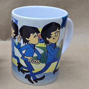 May include: A white ceramic mug featuring a colorful graphic illustration of three cartoon figures with dark hair, wearing blue suits and black boots, walking in a line against a background of green grass and a blue sky with clouds.