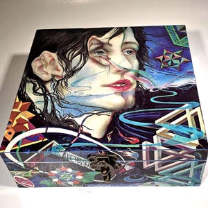 May include: A wooden box with a colorful, abstract design featuring a woman's face, geometric shapes, and swirling lines. The box has a metal latch.