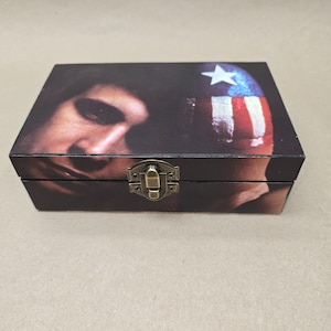 May include: A rectangular wooden box with a hinged lid and a metal clasp. The box features a printed image of a person's face and a painted design of a star and stripes. The color scheme includes shades of brown, red, white, and blue.