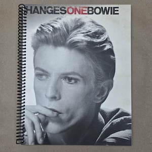 May include: A spiral-bound notebook featuring a black and white portrait of David Bowie. The cover displays the text "CHANGESONEBOWIE" in black and red. The image is a close-up of Bowie's face, with his hand near his mouth.