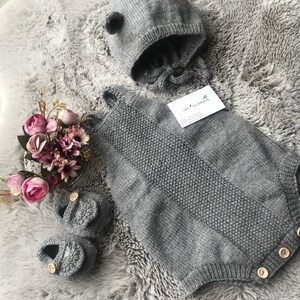May include: A grey knitted baby outfit with a matching hat and booties. The outfit has a sleeveless bodice and long legs. The hat has a small pom-pom on top. The booties have wooden buttons.
