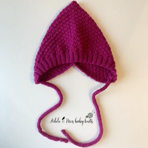 May include: A pink knitted baby bonnet with a pointy hood and ties. The bonnet is made with a textured stitch and has a simple design.