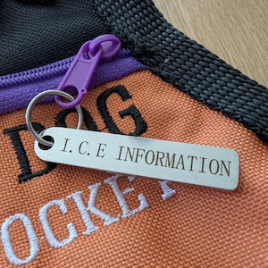 Op de afbeelding: Een zilveren metalen tag met de tekst "I.C.E INFORMATION" bevestigd aan een sleutelhanger. De tag bevindt zich naast een oranje en zwarte hondenpocket met wit en zwart geborduurde tekst.