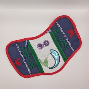 May include: A white fabric pouch with a red, green, and blue trim. The pouch has two zippers and features the text "Adopt Me" with a mermaid, crab, and seashells embroidered on it.