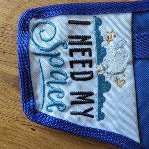 K&ouml;nnte beinhalten: Eine wei&szlig;e Stofftasche mit blauem Besatz und dem gestickten Text "I need my Space" in schwarzem und blauem Garn. Die Tasche hat einen blauen Trageriemen.