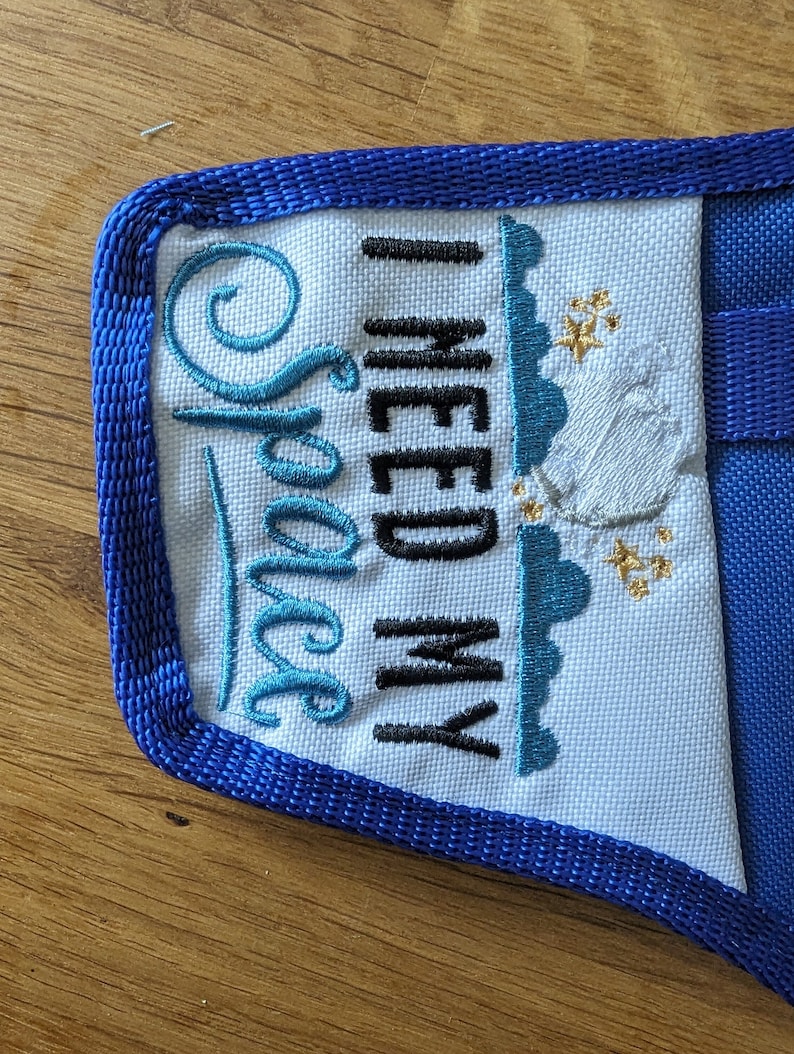 K&ouml;nnte beinhalten: Eine wei&szlig;e Stofftasche mit blauem Besatz und dem gestickten Text "I need my Space" in schwarzem und blauem Garn. Die Tasche hat einen blauen Trageriemen.