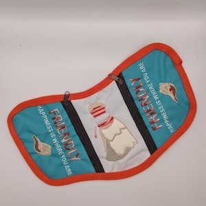 May include: A turquoise blue pouch with a white and red embroidered design of a woman in a dress. The pouch has two zippers and the text "FRIENDLY HAPPINESS IS WHERE YOU ARE" is embroidered on both sides.