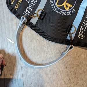 May include: A black nylon dog training bag with a grey strap and two metal D-rings. The bag has the text "OFFICIAL DOG GEAR" and a logo with the letters "DG" in gold.