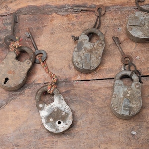 May include: A collection of antique metal padlocks with keys, arranged on a weathered wooden surface. The padlocks are various sizes and shapes, showing signs of age and wear. Some have keys attached with rope or chains.