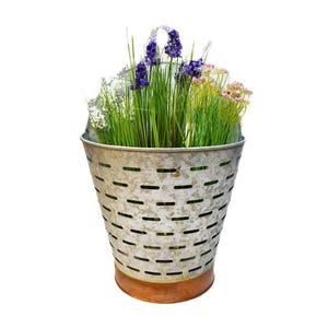 May include: A galvanized metal bucket with a copper-colored base, filled with green grass and colorful flowers. The bucket has a handle and a series of horizontal slots. The flowers include purple, white, and pink blooms.