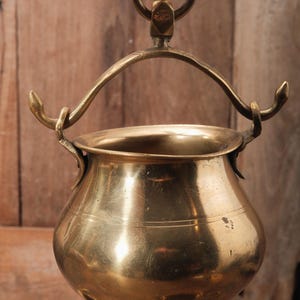 May include: A vintage brass pot with a rounded body and a curved handle. The pot has a small loop at the top for hanging and two decorative hooks on the sides. The brass has a warm, golden hue and shows signs of age.