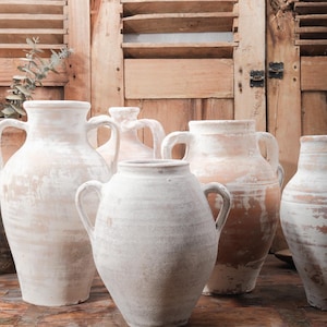 May include: A collection of antique, off-white ceramic vases with handles. The vases vary in size and shape, with a distressed, textured finish. The background features weathered wooden shutters and a rustic wooden surface, creating a vintage aesthetic.