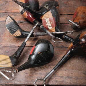 May include: A collection of vintage golf club bottle openers. The openers feature wooden club heads in various colors, including black, brown, and red, with metal bottle opener attachments. The clubs are arranged on a rustic wooden surface.