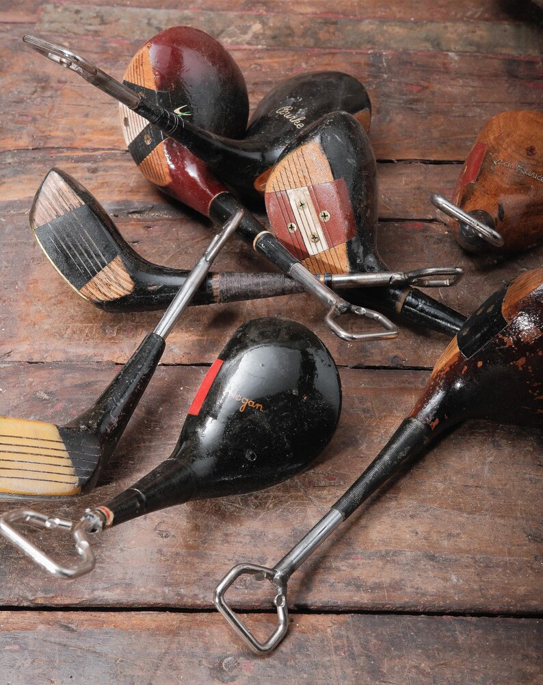 May include: A collection of vintage golf club bottle openers. The openers feature wooden club heads in various colors, including black, brown, and red, with metal bottle opener attachments. The clubs are arranged on a rustic wooden surface.