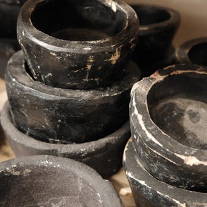 May include: A stack of several black, round, wooden bowls with a distressed finish. The bowls have a dark, matte appearance with visible wear and tear, and some areas of exposed wood.