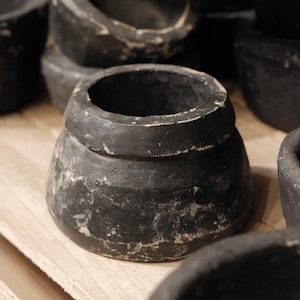 May include: A collection of black, handcrafted, small, round, wooden bowls with a distressed finish. The bowls have a wide opening and a slightly textured surface. The bowls are arranged on a light-colored wooden surface.