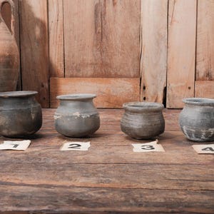 Peut inclure: Quatre petits pots en c&eacute;ramique grise et patin&eacute;e, de forme arrondie et &agrave; large ouverture, sont dispos&eacute;s sur une surface en bois. Chaque pot est num&eacute;rot&eacute; sur une petite &eacute;tiquette. Un grand pichet vieilli est visible en arri&egrave;re-plan.