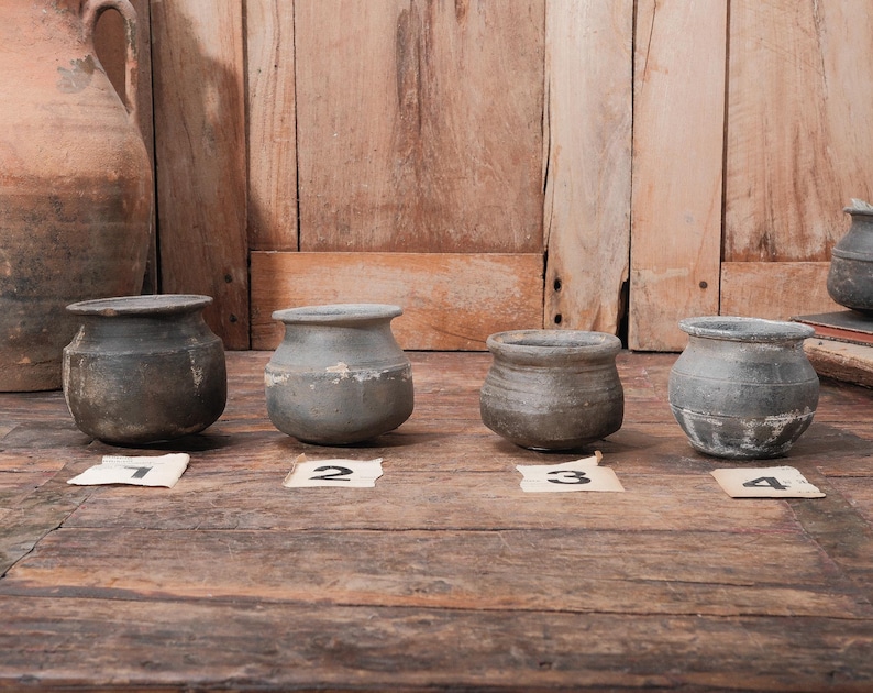 Peut inclure: Quatre petits pots en c&eacute;ramique grise et patin&eacute;e, de forme arrondie et &agrave; large ouverture, sont dispos&eacute;s sur une surface en bois. Chaque pot est num&eacute;rot&eacute; sur une petite &eacute;tiquette. Un grand pichet vieilli est visible en arri&egrave;re-plan.