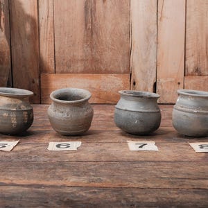 Peut inclure: Quatre petits pots en c&eacute;ramique patin&eacute;s, aux corps arrondis et larges ouvertures, sont expos&eacute;s sur une surface en bois. Chaque pot pr&eacute;sente une texture et une couleur uniques, allant du gris fonc&eacute; au gris clair. Des &eacute;tiquettes num&eacute;rot&eacute;es sont plac&eacute;es devant chaque pot.