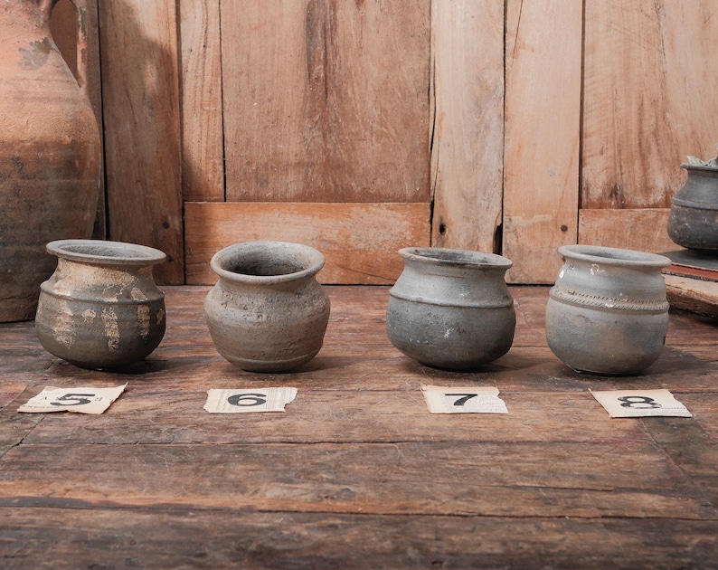 Peut inclure: Quatre petits pots en c&eacute;ramique patin&eacute;s, aux corps arrondis et larges ouvertures, sont expos&eacute;s sur une surface en bois. Chaque pot pr&eacute;sente une texture et une couleur uniques, allant du gris fonc&eacute; au gris clair. Des &eacute;tiquettes num&eacute;rot&eacute;es sont plac&eacute;es devant chaque pot.