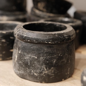 May include: A collection of small, round, black pots with a distressed, aged appearance. The pots have a wide opening and a slightly textured surface. The pots are arranged on a light-colored wooden surface.