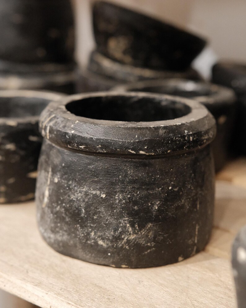 May include: A collection of small, round, black pots with a distressed, aged appearance. The pots have a wide opening and a slightly textured surface. The pots are arranged on a light-colored wooden surface.