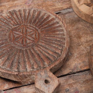 May include: Close-up of several rustic, round wooden serving boards with handles. The boards have intricate carved designs, including a central starburst pattern and radial lines. The wood has a weathered, aged appearance, with variations in color and texture.