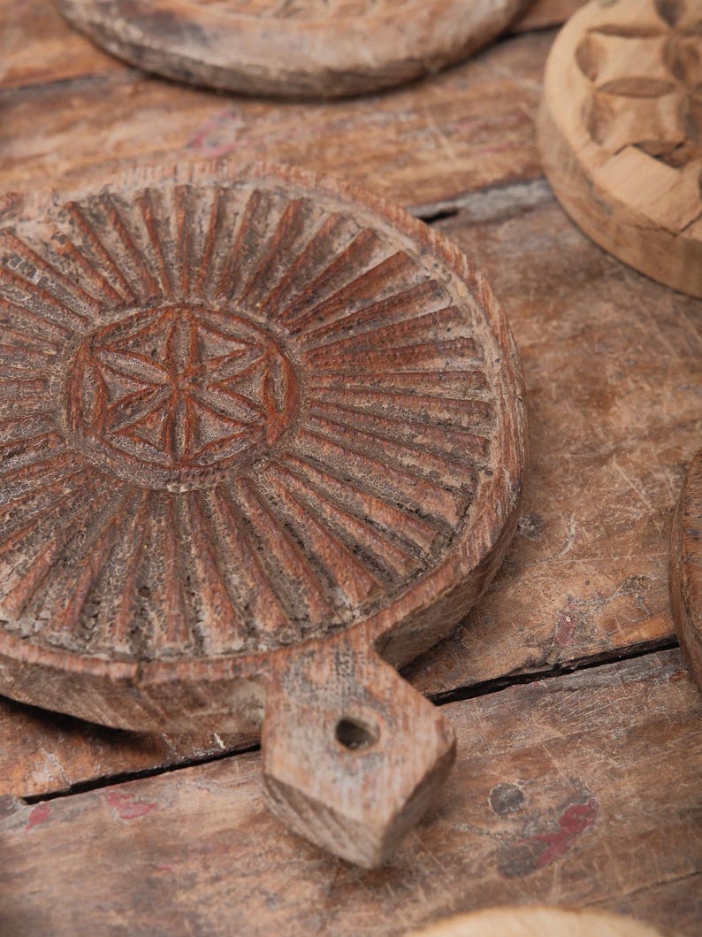May include: Close-up of several rustic, round wooden serving boards with handles. The boards have intricate carved designs, including a central starburst pattern and radial lines. The wood has a weathered, aged appearance, with variations in color and texture.