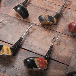 May include: A collection of vintage golf club bottle openers. The openers feature black, yellow, and red accents. The openers are arranged on a weathered wooden surface.